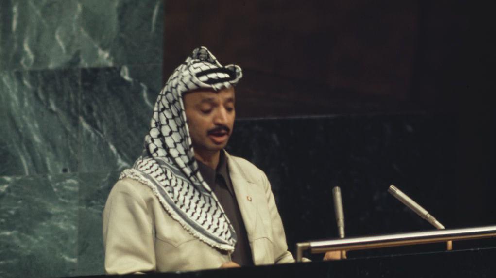 ياسر عرفات، مرتدياً الكوفية الفلسطينية وبزة عسكرية، يلقي خطاباً أمام الجمعية العامة للأمم المتحدة في نيويورك عام 1974