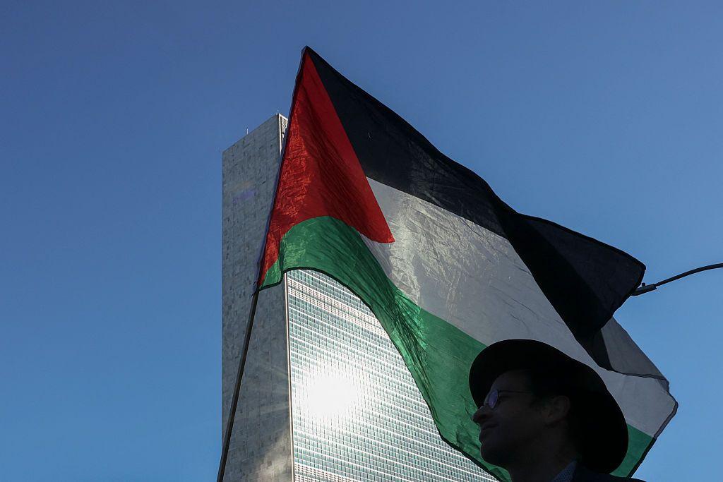 متظاهر يرتدي قبعة سوداء ونظارات تبدو طبية يحمل العلم الفلسطيني وفي خلفية الصورة مقر الأمم المتحدة في بناء شاهق وسماء زرقاء صافية.