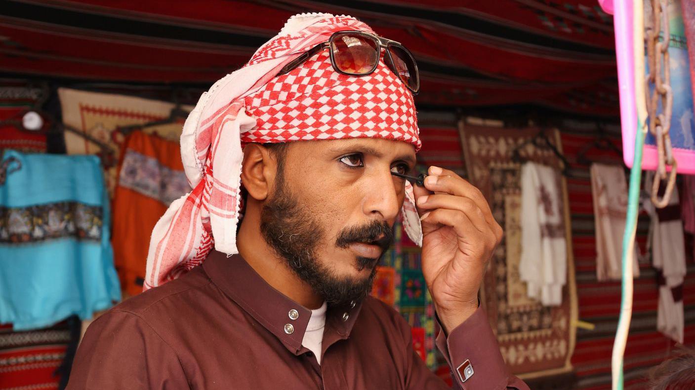 رجل يضع كحل عيون أسود، ويرتدي قميصاً بنياً، ويعتمر كوفية أو غطاء رأس عربي تقليدي باللونين الأحمر والأبيض. 
