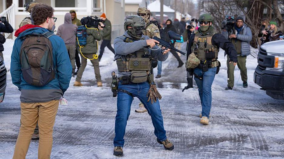 ضابط من هيئة الهجرة والجمارك الأميركية (ICE) (على اليمين) وآخر من الضباط الفيدراليين يحمل جهازاً للتحكم في الحشود (في الوسط) يقفان عند تقاطع في مينيابوليس حيث تجمع المحتجون بعد مقتل رينيه نيكول غود برصاص واحد من عناصر الـICE، بينما يقف المارة والصحفيون داخل التقاطع كذلك.