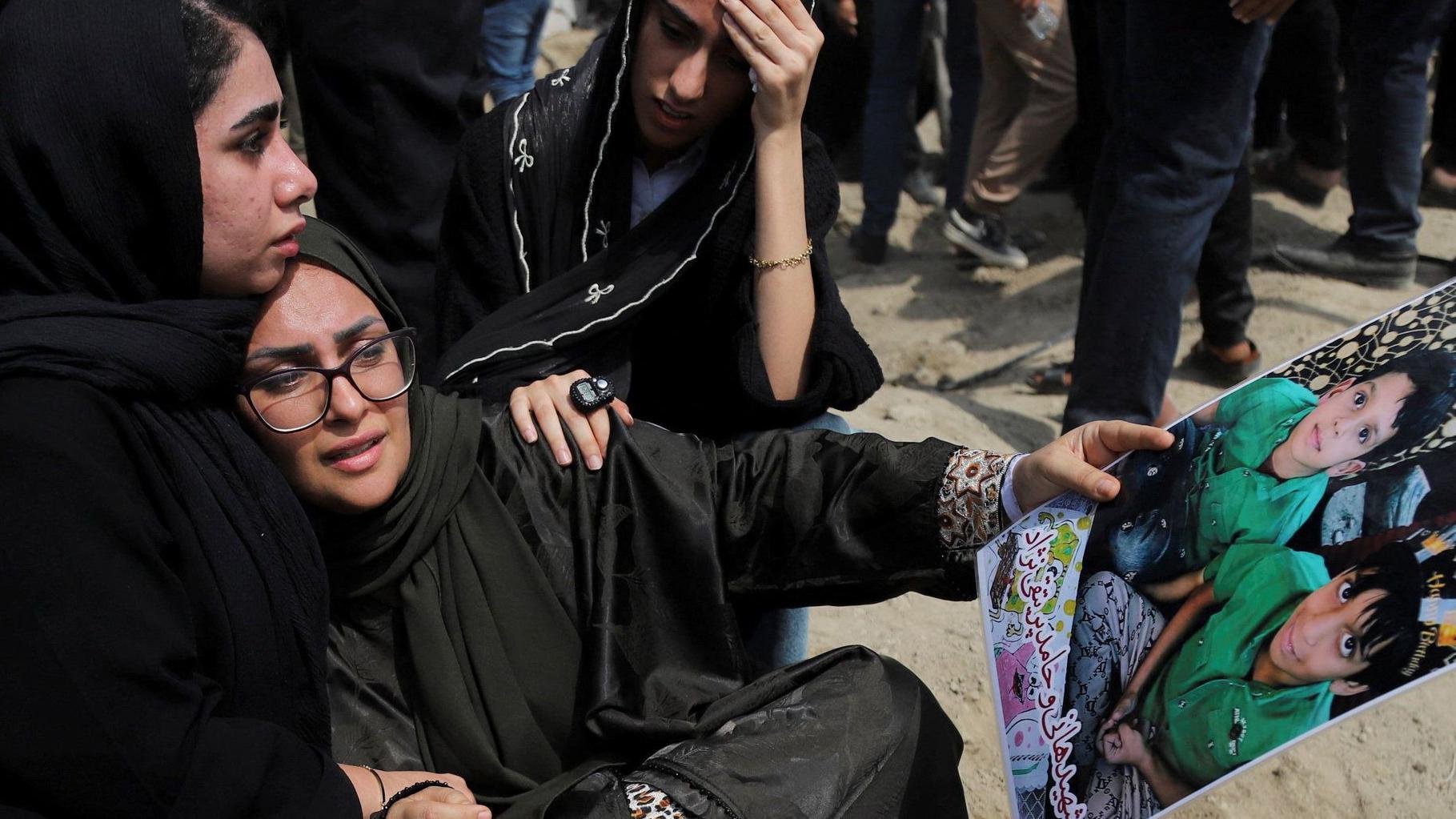 امرأة تحمل صورة لطفلين تجلس على الأرض وتتلقى العزاء من امرأتين أخريين؛ وتظهر أرجل العديد من الأشخاص الآخرين الواقفين حولهم. 