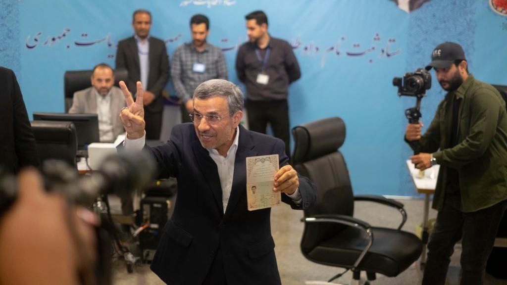 محمود أحمدي نجاد يسجل اسمه في مقر الانتخابات التابع لوزارة الداخلية الإيرانية، ويمسك بيده ورقه ويلوح بيده بعلامة النصر ، وفي الخلف مصور بيده كاميرا يلتقط له صورة.